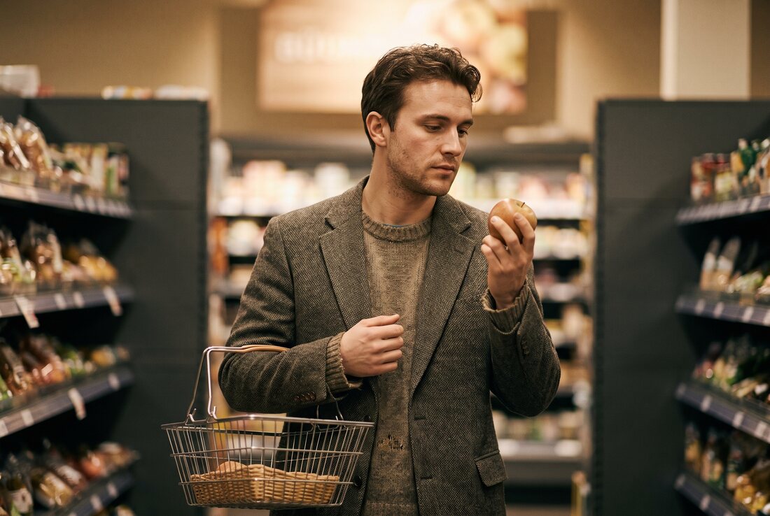 A man pausing in a supermarket aisle with a small basket, weighing up whether to add an apple to it.