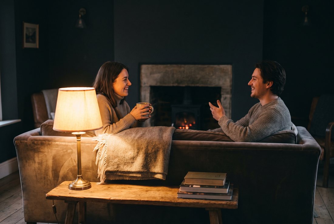 Two people sitting at either end of a sofa in lamplight, talking easily over a mug of tea.